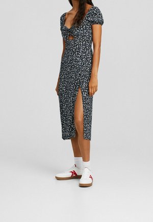 Kobieta w czarnej sukience midi w kwiatowy wzór z bufkami na rękawach, wycięciem z przodu, rozcięciem na boku na udzie, białe skarpetki i białe trampki z czerwonymi akcentami.