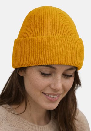 Young woman with long brown hair wearing a mustard yellow knit beanie and beige sweater, looking down with a slight smile.