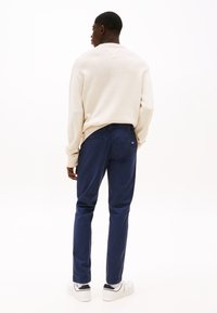 Muž stojaci chrbtom, oblečený v krémovom svetri, tmavomodrých nohaviciach a bielych teniskách s čiernym detailom na päte, na bielom pozadí.