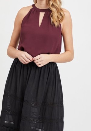 Femme portant un haut sans manches bordeaux style halter avec une encolure goutte et une jupe noire taille haute avec des panneaux en dentelle.