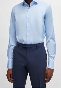 Camisa de vestir azul claro de manga larga, con cuello abotonado y textura suave, combinada con pantalones azul oscuro que presentan un patrón sutil.