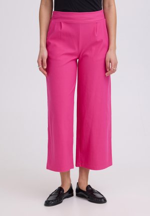 Žena nosí jasnoružové široké skrátené nohavice s čiernymi mokasínami, stojaca pred jednofarebným svetlým pozadím.