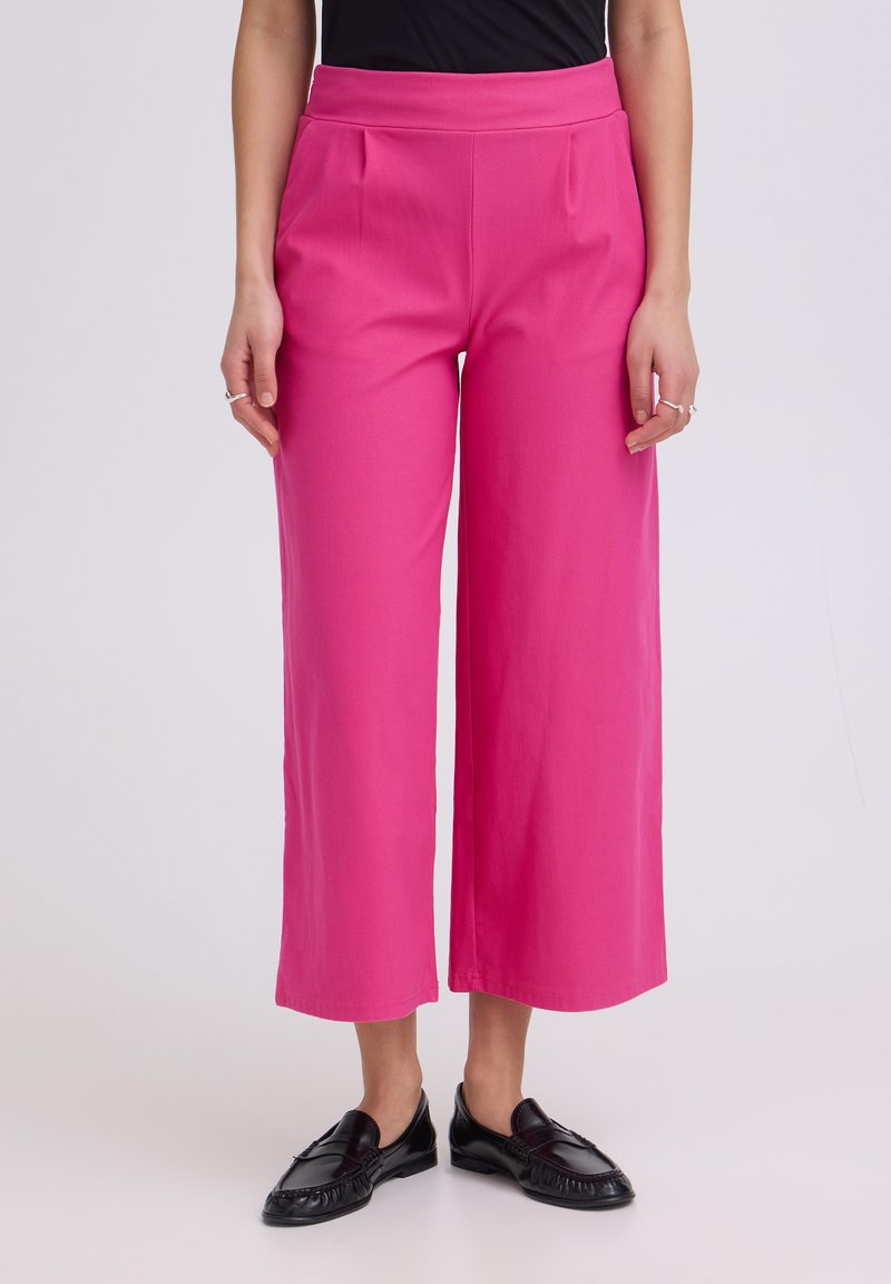 Femme portant un pantalon large court rose vif avec des mocassins noirs, debout devant un fond clair uni.