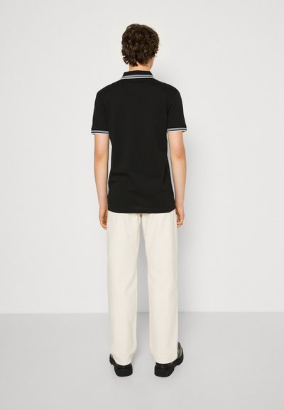 Polo shirt noir avec col et poignets rayés blancs, associé à un pantalon large crème et des chaussures noires. Design minimal.