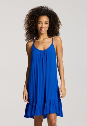 Žena s kučeravými vlasmi, ktorá má na sebe modré šaty bez rukávov s tenkými ramienkami a náhrdelník z korálikov, usmieva sa do kamery.