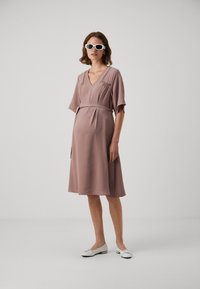 Mauve midi sukienka z dekoltem w kształcie litery V, luźnymi rękawami i przewiązanym w pasie. Gładka faktura, noszona z białymi okularami przeciwsłonecznymi i płaskimi butami.