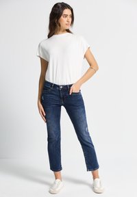 Top blanco de manga corta combinado con jeans de color azul oscuro. Los jeans tienen un ligero desgaste y el modelo usa zapatillas deportivas blancas. Diseño simple y contemporáneo.