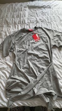 Sivá tričko s krátkym rukávom vyrobené z mäkkej látky s okrúhlym výstrihom a detailom skrúteného predného dielu. Obsahuje červenú značkovú etiketu.