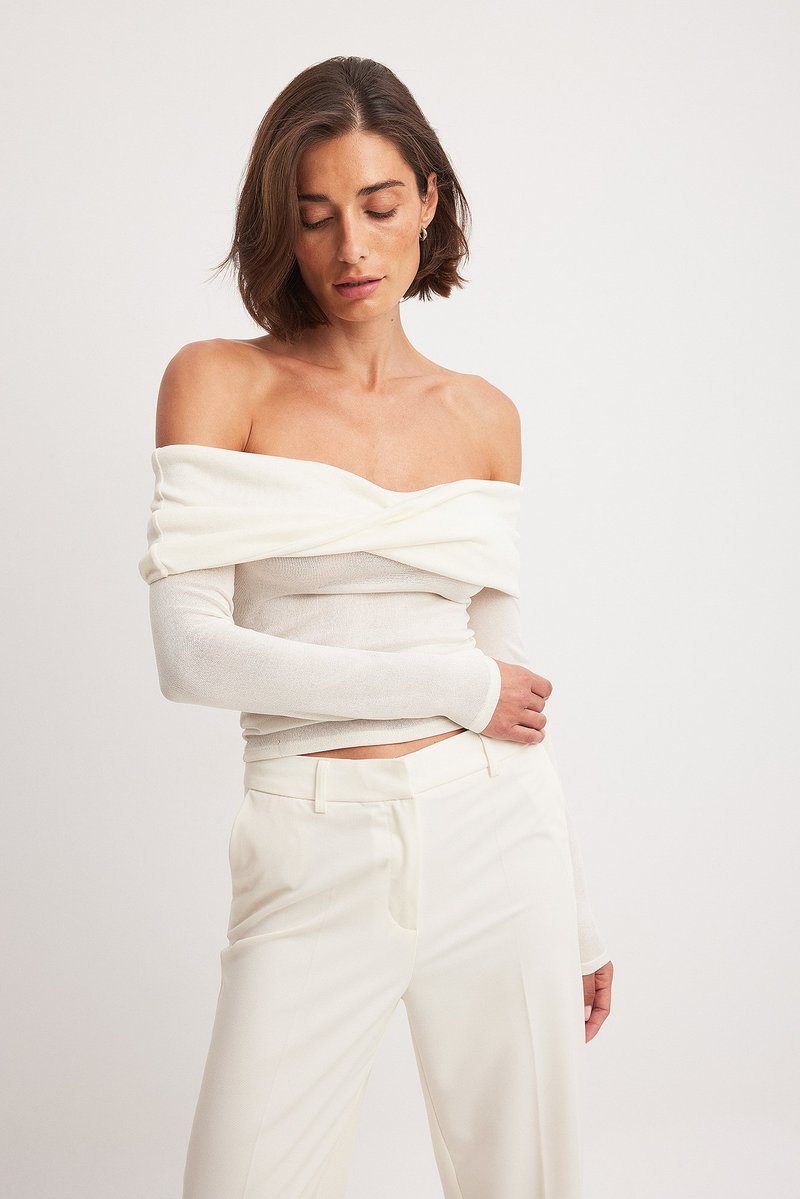 Off-the-shoulder hvid bluse med lange ærmer, tekstureret stof og en snoet design detalje, parret med hvide skræddersyede bukser.