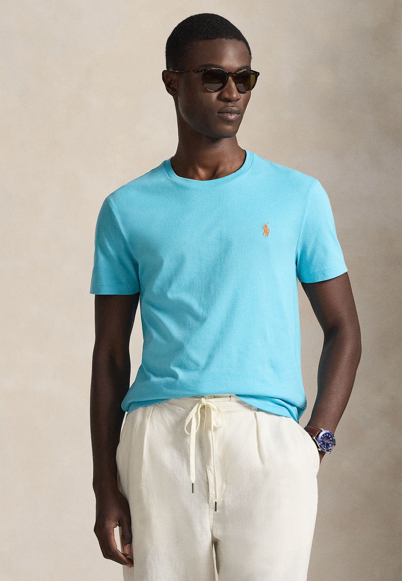 Ανοιχτό γαλάζιο κοντομάνικο μπλου ζάκι με στρογγυλεμένο λαιμόκοψη και μικρό πορτοκαλί λογότυπο. Φορεμένο με ανοιχτό μπεζ παντελόνι και σκούρα γυαλιά ηλίου.