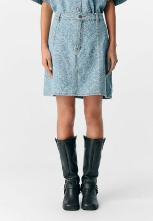 Jupe en denim bleu clair avec un motif cachemire, dotée d'une fermeture à boutons et de passants de ceinture. Associée à des bottes en cuir noires hautes.