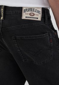 Schwarze Denim-Jeans mit einem Standard-Schnitt, strukturiertem Finish und geschwungenen Nähten an den hinteren Taschen. Das Etikett trägt die Aufschrift "REPLAY BLUE JEANS."