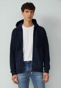 Marinefarbener Strick-Zip-Hoodie mit strukturiertem Oberteil, zwei frontalen Taschen und einem einfarbigen unteren Abschnitt. Kombiniert mit einem weißen T-Shirt und Jeans.