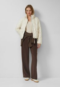 Weiße gesteppten Jacke mit hohem Kragen, kombiniert mit einem beigen Pullover und braunen, weit geschnittenen Hosen, abgerundet durch cremefarbene Slip-On-Schuhe.