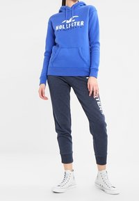 Blå hoodie med en framficka och vit "Hollister California" logotyp, tillsammans med mörkgrå sweatpants med vit branding och mudd. 