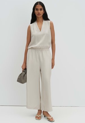 Frau trägt einen ärmellosen beigen Overall mit weitem Bein, hält eine kleine graue Tasche in der Hand und trägt beigefarbene Sandalen mit Absatz.