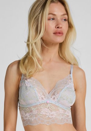 Blonde vrouw die een pastelgekleurde kanten bralette draagt met bloemmotieven, dunne bandjes en een kleine roze roos als detail in het midden.