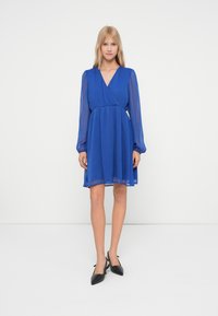 Femeie stă în fața unui fundal alb purtând o rochie albastră regală, până la genunchi, cu mâneci lungi transparente și pantofi slingback negri, cu vârful ascuțit.