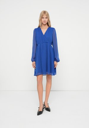 Kvinne står mot en hvit bakgrunn iført en knelengde kongelig blå kjole med gjennomsiktige lange ermer og svarte spisse slingback hæler.