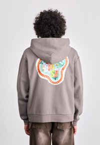 Grey sweatshirt featuring a large colorful embroidered design on the back, with a round shape and fringed edges. Soft fabric texture.