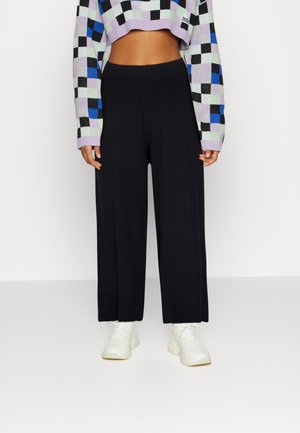 Black wide-leg trousers made of ribbed fabric, paired with a cropped, multicolored checkered sweater, featuring pastel shades and a black accent.