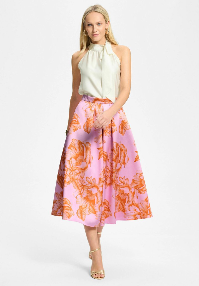 Femme portant un haut écru sans manches à col halter, associé à une jupe midi rose ornée de grandes fleurs orange, et des talons à lanières dorées.