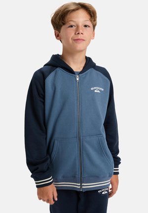 Kapuzen-Zip-Up-Sweatshirt aus weichem Stoff, mit blauem Körper und marineblauen Ärmeln, weißen Akzenten und Fronttaschen. Quiksilver-Logo sichtbar.