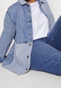 Osoba ubrana w białą koszulkę, dwukolorową dżinsową kurtkę i ciemnoniebieskie dżinsy, siedząca z jedną nogą zgiętą na jasnoszarym podłożu.