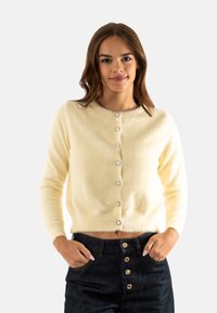 Cárdigan amarillo esponjoso con un escote redondeado, cierre frontal abotonado con botones en tono plateado y mangas largas. Lleva puesto sobre unos jeans oscuros.