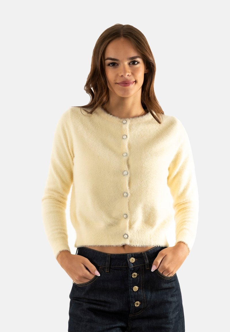 Cárdigan amarillo esponjoso con un escote redondeado, cierre frontal abotonado con botones en tono plateado y mangas largas. Lleva puesto sobre unos jeans oscuros.