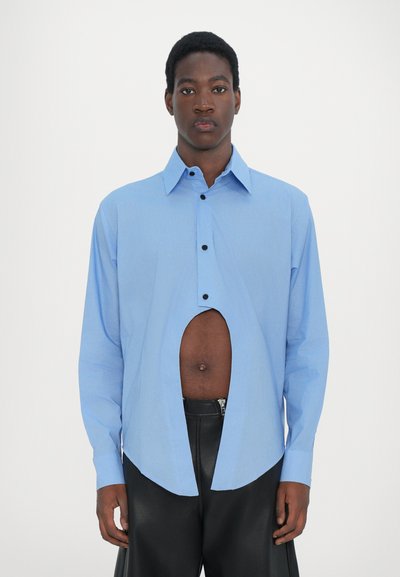 Chemise bleu clair à manches longues avec une grande découpe à la taille, dotée d'un devant à boutons et d'un col pointu, portée sur un pantalon noir.