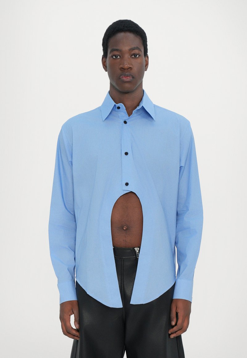 Chemise bleu clair à manches longues avec une grande découpe à la taille, dotée d'un devant à boutons et d'un col pointu, portée sur un pantalon noir.