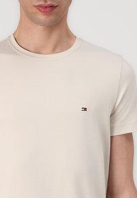 Bežinis medvilninis t-shertas trumpomis rankovėmis, su apvaliu iškirpimu ir mažai išsiuvinėta logotipu, kuriame yra raudonų, baltų ir mėlynų akcentų ant krūtinės.