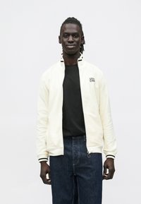 Lys cremefarvet lynlåstrøje med ribbet krave og manchetter, kombineret med en sort t-shirt og mørke jeans. Har diskret branding på brystet.