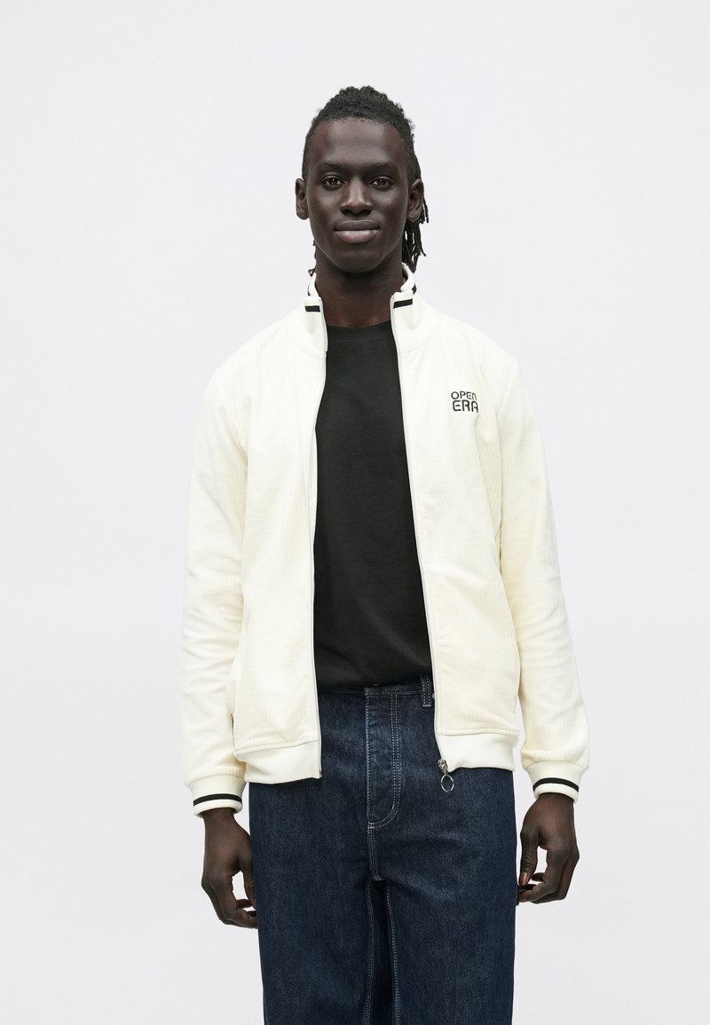 Lys cremefarvet lynlåstrøje med ribbet krave og manchetter, kombineret med en sort t-shirt og mørke jeans. Har diskret branding på brystet.