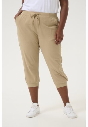 Kvinde iført beige cropped bukser med snor og lommer samt hvide sneakers, står med den ene hånd i lommen.