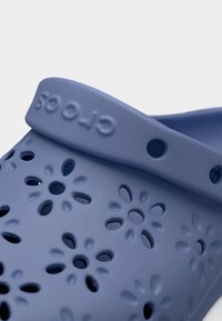 Κοντινό πλάνο μπλε παπουτσιού Crocs με οπές αερισμού σε σχήμα λουλουδιού και ρυθμιζόμενο ιμάντα στη φτέρνα με ανάγλυφο το όνομα της μάρκας.