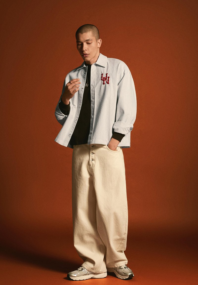 Mann trägt karierte helle Jacke mit rotem Logo über schwarzem Hemd, weite beige Hose und silberne Sneakers, steht vor braunem Hintergrund.