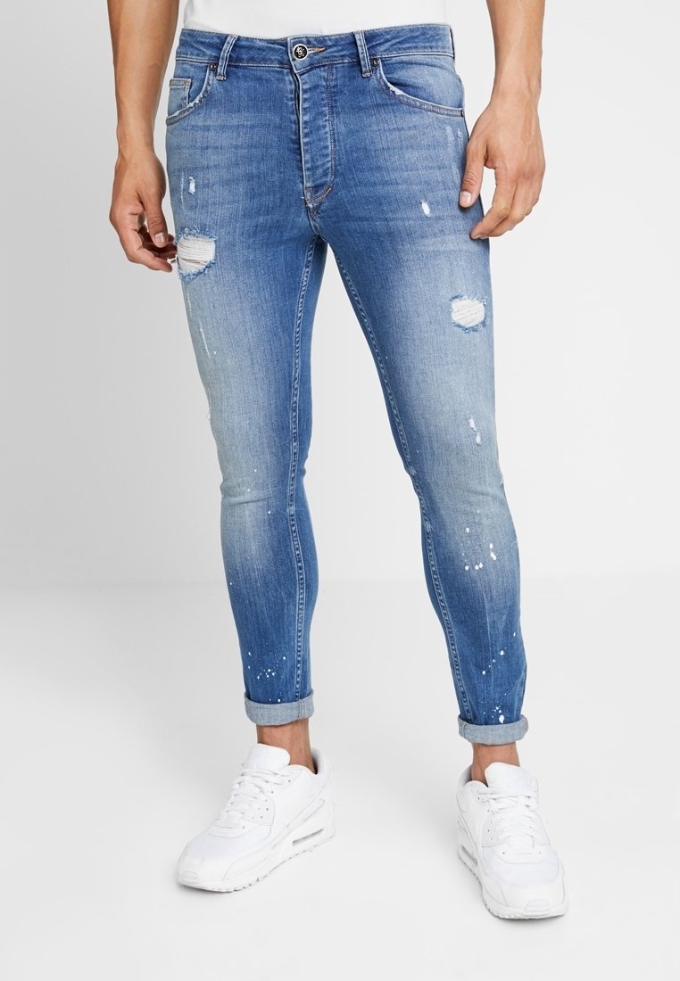 Jeans ajustados de color azul con desgastes y rotos, lavado claro y detalles de manchas de pintura, que cuentan con un diseño estándar de cinco bolsillos y puños enrollados.