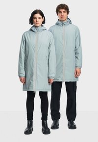 Giacche impermeabili lunghe di colore azzurro chiaro, con cappucci, zip complete e polsini elasticizzati, indossate sopra leggings neri e stivali. Design minimalista, senza fantasie.