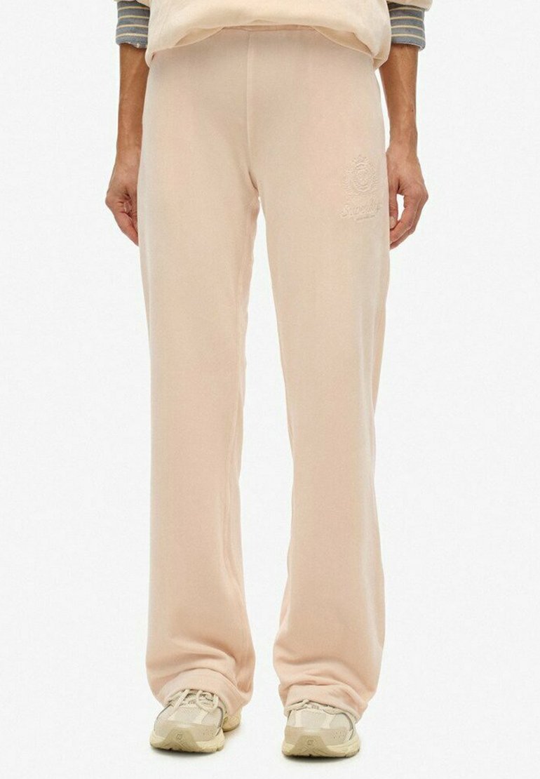 Persoon in beige rechttoe rechtaan sweatpants en bijpassende sneakers, staand met ontspannen handen langs de zijkanten tegen een effen achtergrond.