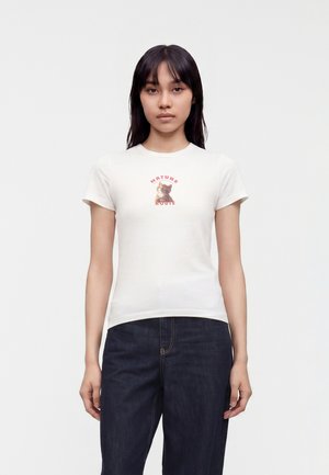 Kvinde med langt mørkt hår iført en hvid T-shirt med en killing og ordene "Nature Route" og mørkeblå jeans, stående mod en ensfarvet baggrund.