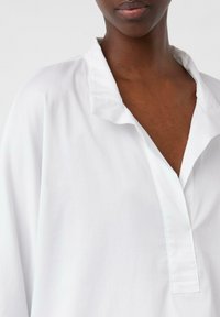 White collared shirt with a deep V-neck and relaxed fit. Smooth texture and minimal seams, featuring a subtle ruffle at the collar.