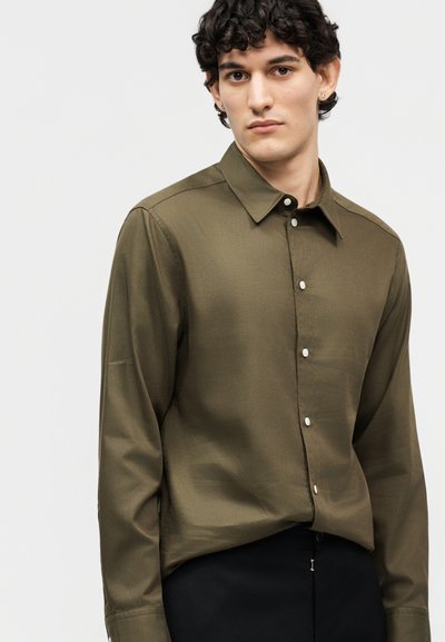 Νεαρός άνδρας με κατσαρά μαλλιά, φορώντας πουκάμισο με μακριά μανίκια, ελιάς πράσινο, κουμπωμένο και μαύρο παντελόνι, σε απλό ανοιχτό φόντο.