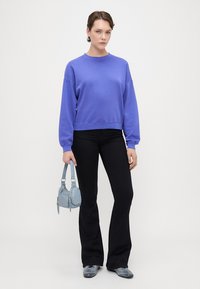Sudadera morada con cuello redondo y hombros caídos, combinada con pantalones negros de campana y un pequeño bolso azul claro.