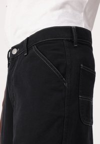 Svarta denimjeans med kontrasterande vit sömnad. Har knäppning i midjan, snedställda framfickor och en bakficka på höger sida.
