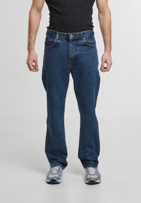 Jeans din denim albastru închis cu un design clasic drept, cinci buzunare și închidere cu nasture, purtate cu adidași gri.