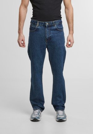 Mørkeblå denimjeans med klassisk rett ben-design, fem lommer og knappeåpning, iført grå treningssko.