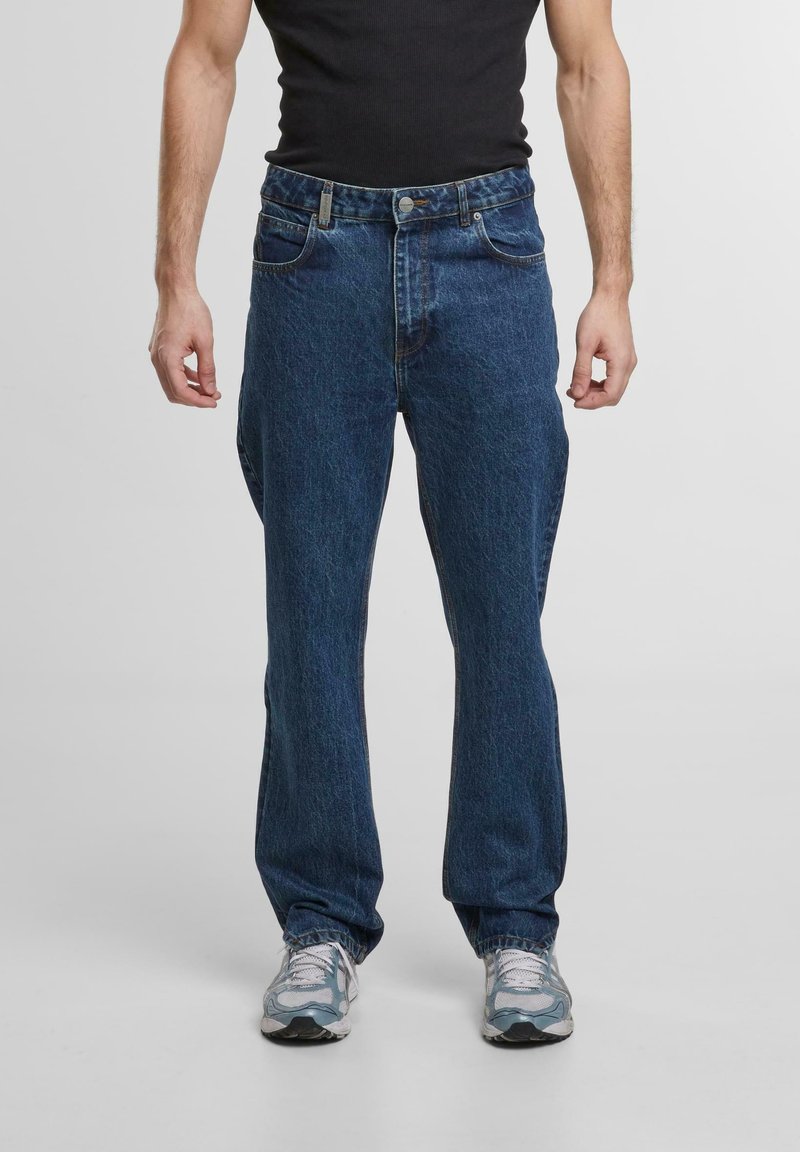 Jeans din denim albastru închis cu un design clasic drept, cinci buzunare și închidere cu nasture, purtate cu adidași gri.