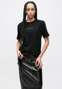 Černé bavlněné tričko s krátkými rukávy a stříbrným nápisem "PINKO". Kombinováno s dlouhou lesklou černou koženou sukní vyznačující se hladkou texturou.